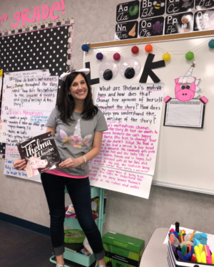 Marine with a unicorn headband for unicorn day, standing in front of the anchor chart holding the book, Thelma the Unicorn.