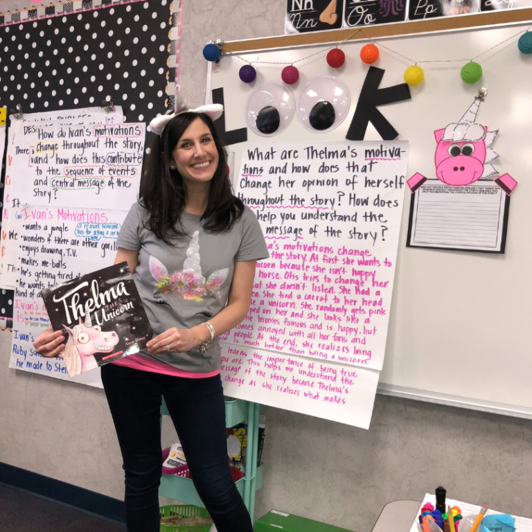 Marine with a unicorn headband for unicorn day, standing in front of the anchor chart holding the book, Thelma the Unicorn.